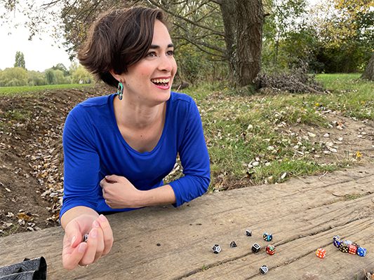 Caitlin, a young white woman with dark brown hair, wears a blue shirt and is seated at a wood table. She is smiling and rolling dice onto the table.